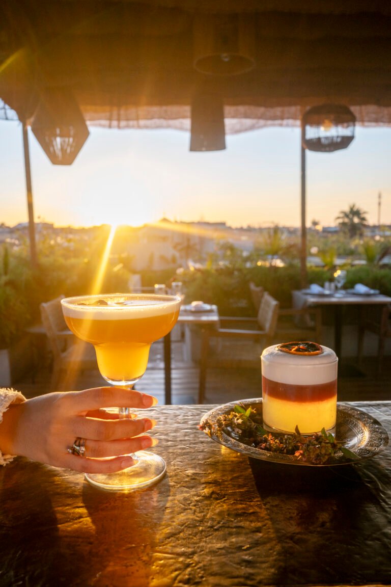 A hand holding a golden cocktail at sunset on the Bidaya rooftop terrace in Marrakech, with a second layered drink on a brass tray in the background
