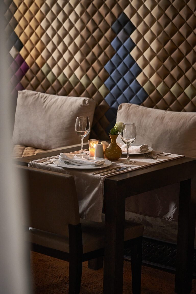A candlelit dinner table for two at Almaha Marrakech, featuring elegant glassware and a yellow rose vase against a geometric silk-tiled wall