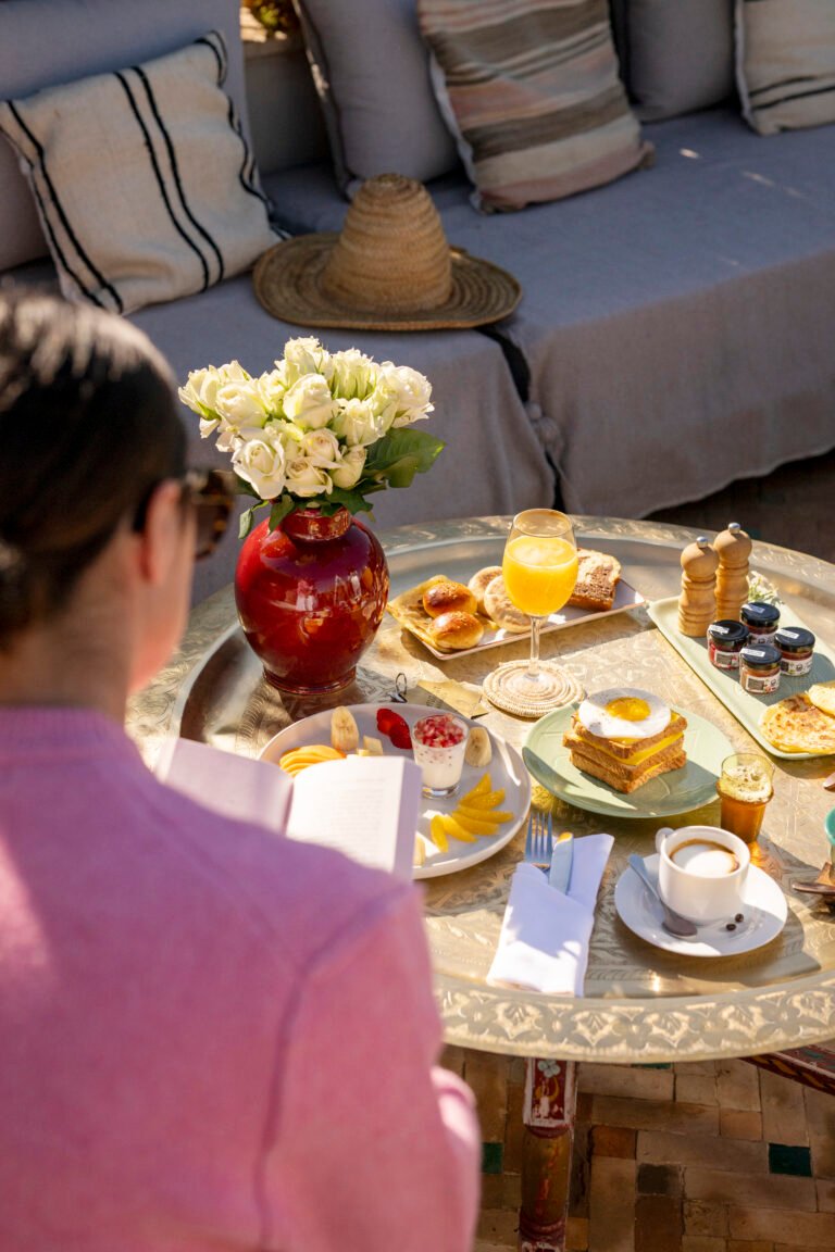 A guest reading a book while enjoying a luxury Moroccan breakfast on a sunlit terrace at Almaha Marrakech, featuring fresh fruit, pastries, and orange juice on a brass tray.