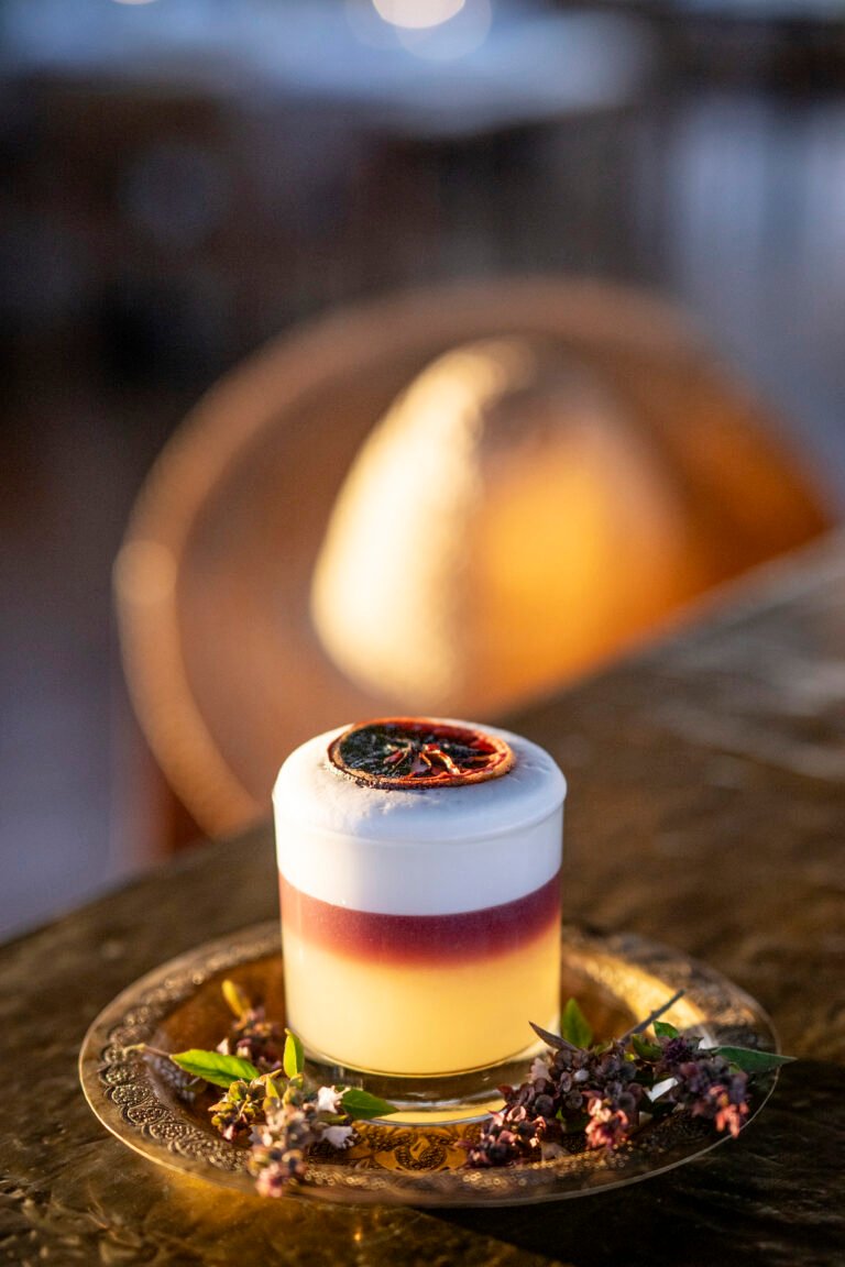 A layered craft cocktail with a thick foam head and dried citrus garnish, served on a traditional brass plate at Almaha Marrakech