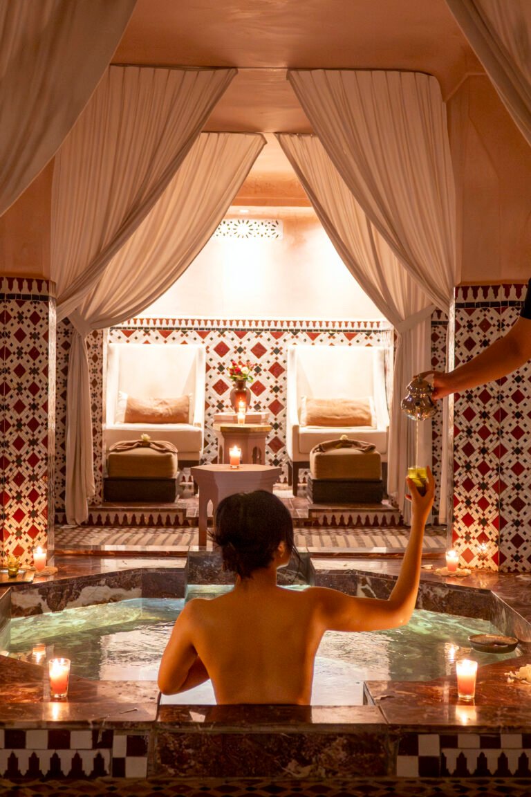 A guest relaxing in an indoor marble pool at Almaha Marrakech spa, being served traditional Moroccan mint tea in a candlelit atmosphere.