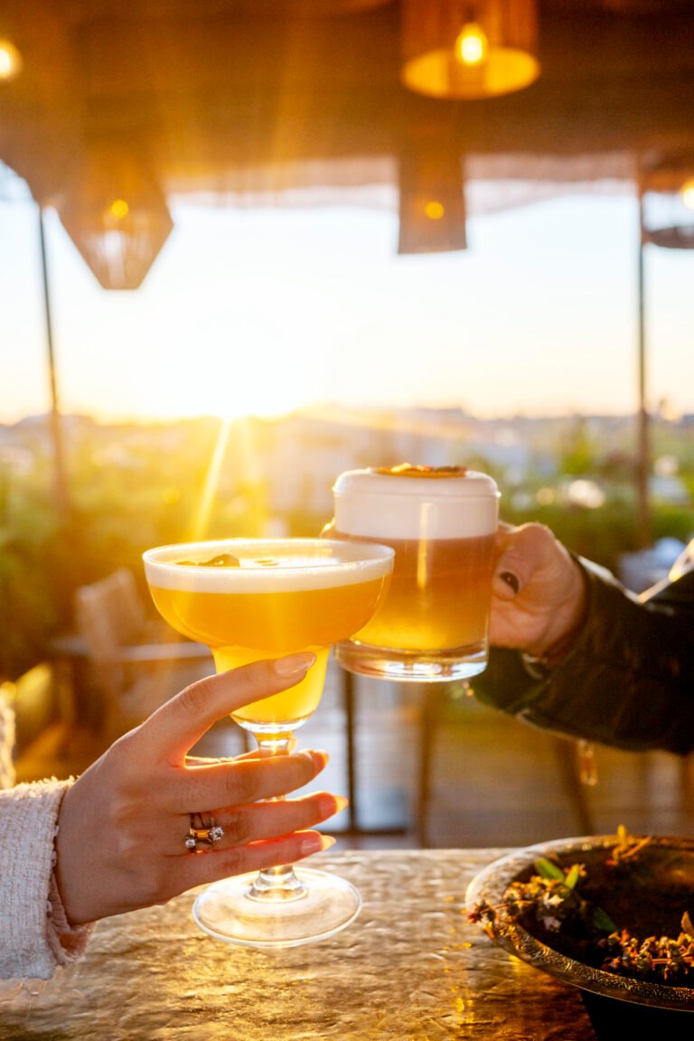 Two guests toasting with golden sunset cocktails on the Bidaya Rooftop at Almaha Marrakech, overlooking the city at dusk.