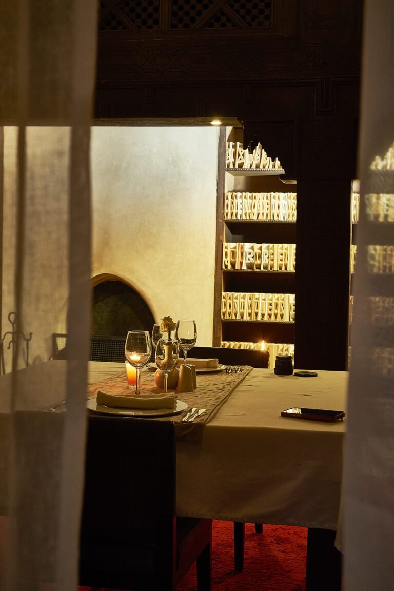 An elegant candlelit dinner table set for two in The Library at Almaha Marrakech, seen through soft curtains with illuminated book sculptures in the background