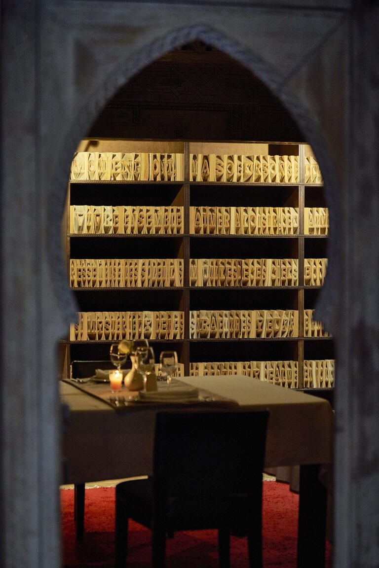 View through a Moroccan archway of an illuminated book sculpture wall by Charles Kaisin in The Library restaurant at Almaha Marrakech