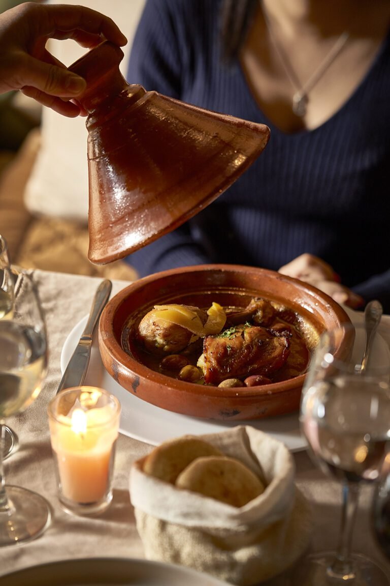 A waiter lifting the lid of a traditional clay tajine to reveal a lemon and olive chicken dish at Almaha Marrakech restaurant